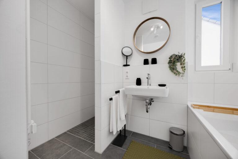 Modernes, helles Badezimmer mit Wand-WC, weißem Waschbecken, Spiegel, begehbarer Dusche und dekorativen Elementen für ein stilvolles Ambiente