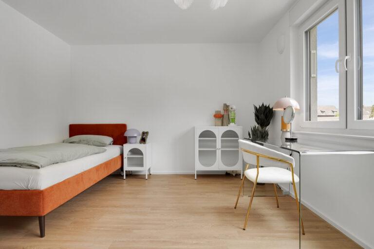 Modern eingerichtetes Gästezimmer mit orangefarbenem Einzelbett, stilvollem Arbeitsbereich am Fenster und Designermöbeln auf hellem Holzboden