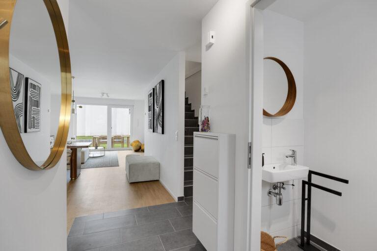 Heller moderner Flur mit großem runden goldenen Spiegel, Blick ins Wohnzimmer und Zugang zum Gäste-WC mit kleinem Waschbecken