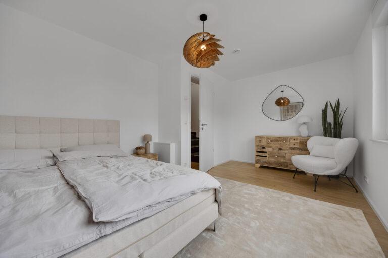 Modern eingerichtetes Schlafzimmer mit großem Bett, Holzkommode, Designlampe, gemütlichem Sessel und hellen Farbtönen für ein stilvolles Wohnambiente