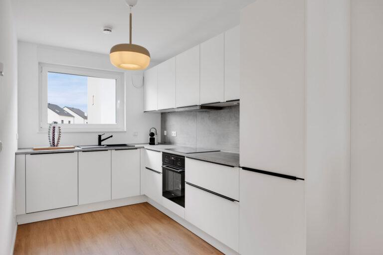 Moderne weiße Movida Einbauküche in L-Form mit Holzboden, grauer Arbeitsplatte und Blick aus dem Fenster auf Baustelle