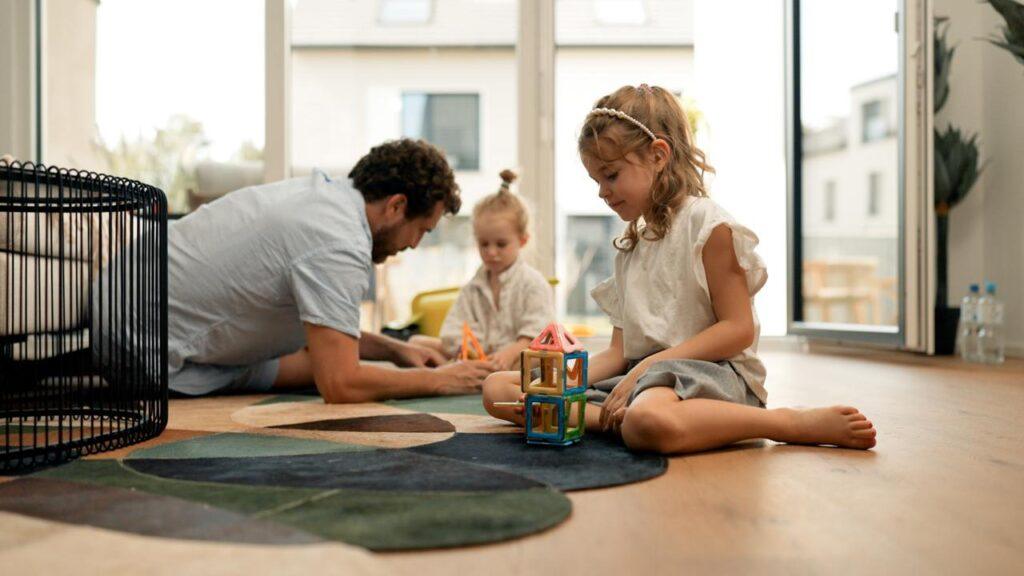 Kinder spielen mit Bausteinen auf dem Wohnzimmerboden, während der Vater mit ihnen spielt, in einer Movida-Immobilie zur Miete.