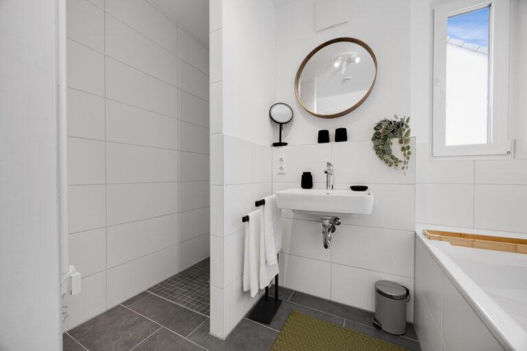 Modernes, helles Badezimmer mit Wand-WC, weißem Waschbecken, Spiegel, begehbarer Dusche und dekorativen Elementen für ein stilvolles Ambiente