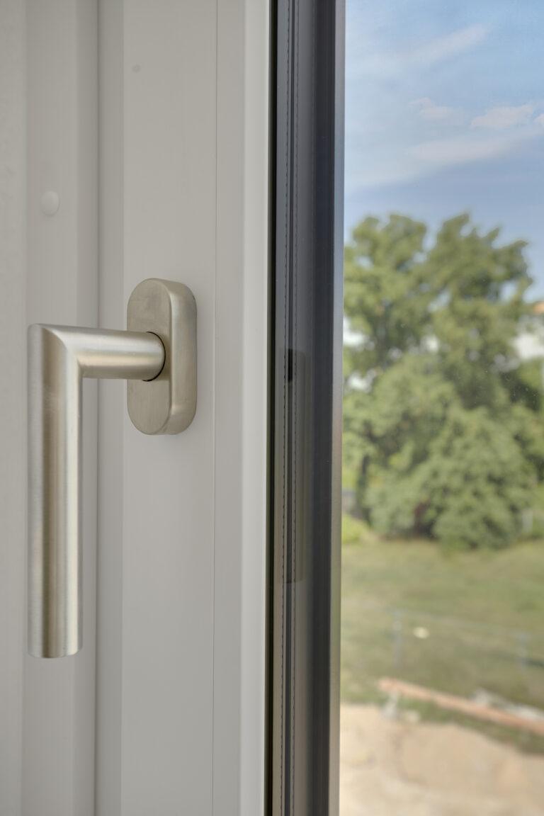 Nahaufnahme eines modernen Edelstahl - Fenstergriffs an einem weißen Kunststofffenster mit Blick ins Grüne