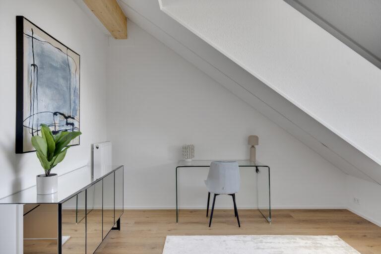 Modernes Arbeitszimmer im Dachgeschoss mit Schreibtisch, Stuhl und dekorativen Elementen in einer Movida-Immobilie zur Miete.