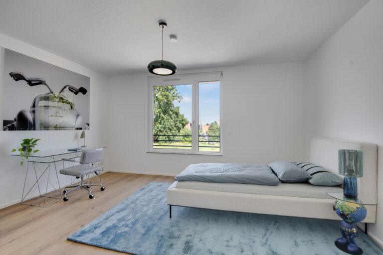 Geräumiges Schlafzimmer mit großem Fenster, Schreibtisch und modernem Design in einer Movida-Immobilie zur Miete