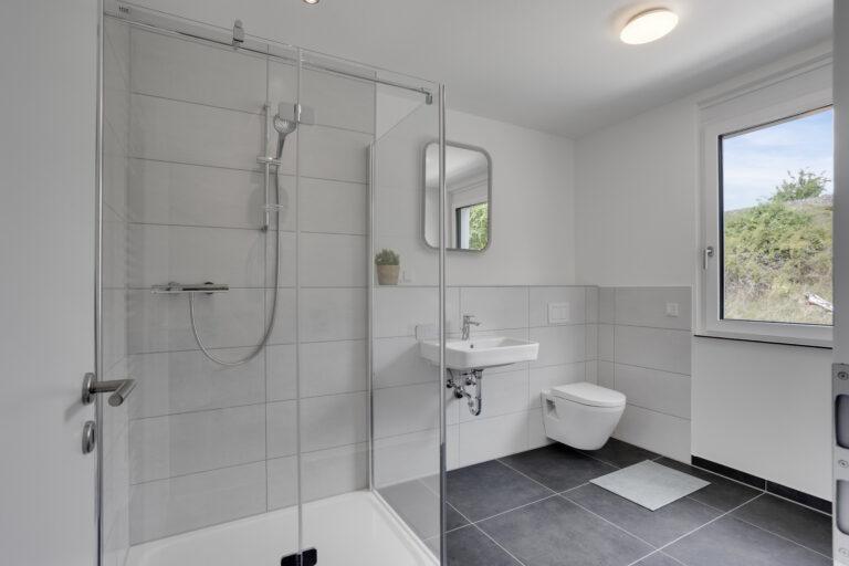 Modernes Badezimmer mit bodengleicher Dusche, großem Fenster und grauen Fliesen im MoVida Haus