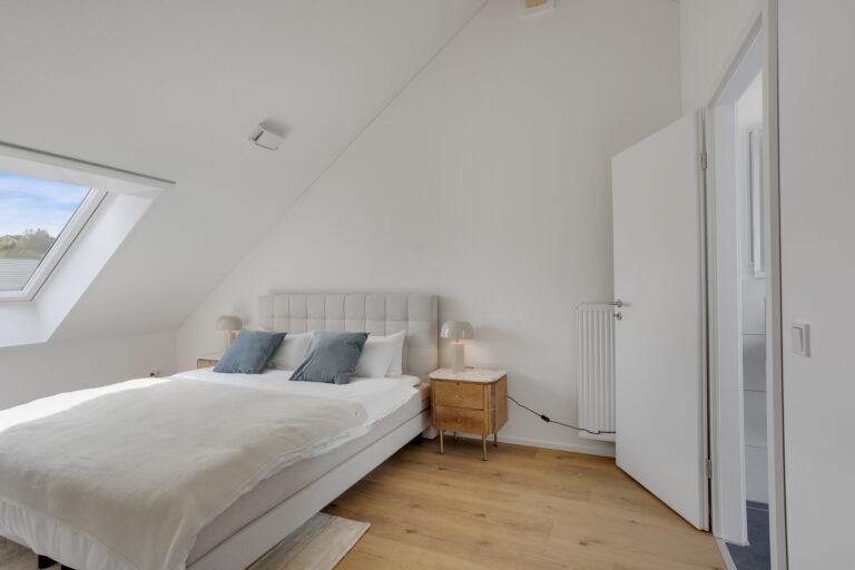 Modernes Schlafzimmer im Dachgeschoss mit schräger Decke, großem Dachfenster, Doppelbett mit hellgrauem Kopfteil, zwei Nachttischen aus Holz und heller, minimalistischer Einrichtung.