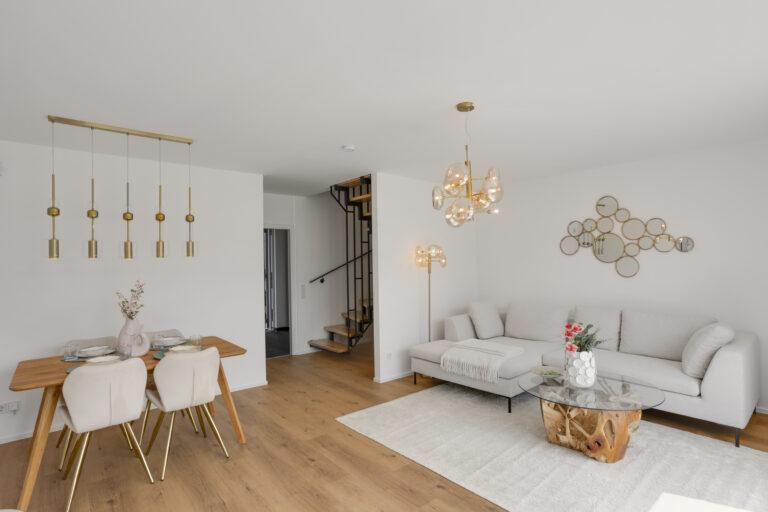 Modern eingerichtetes Wohnzimmer mit heller Couch, Glas-Couchtisch mit Holzfuß, stilvoller Beleuchtung und offener Treppe, große Fensterfront mit viel Tageslicht.