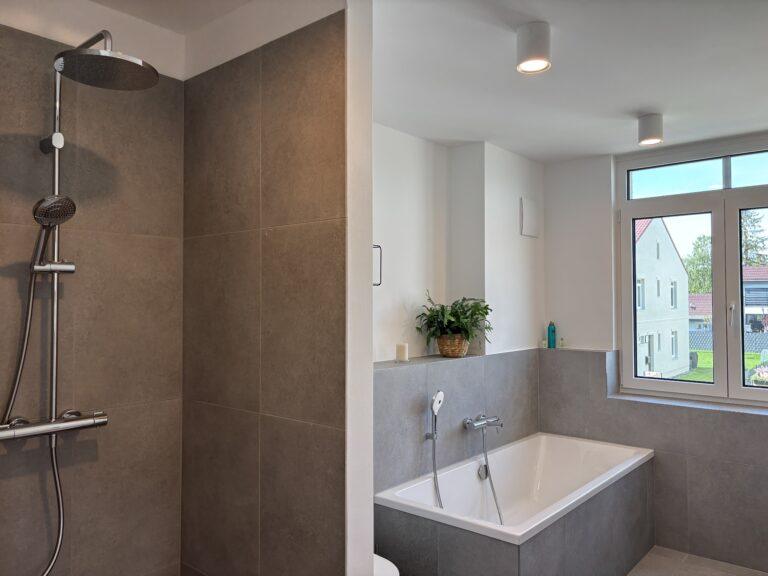 Modernes Badezimmer mit bodengleicher Dusche, Badewanne, großem Fenster und grauen Fliesen im MoVida Haus