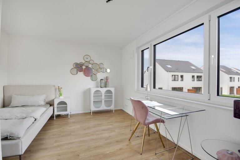 Modern eingerichtetes Gästezimmer mit grauem Einzelbett, stilvollem Arbeitsbereich am Fenster und Designermöbeln auf hellem Holzboden