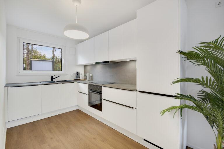 Moderne weiße Movida Einbauküche in L-Form mit Holzboden, grauer Arbeitsplatte und Blick aus dem Fenster