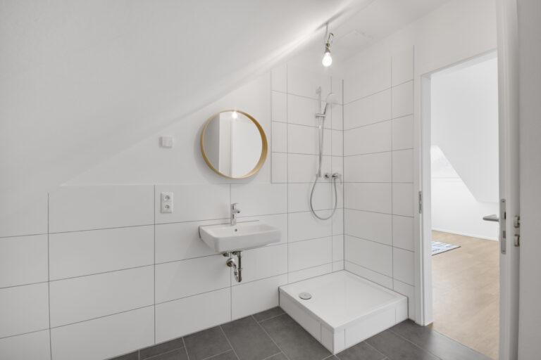 Modernes Badezimmer im Dachgeschoss eines Reihenhauses mit bodengleicher Dusche, weißen Fliesen und Tageslicht.