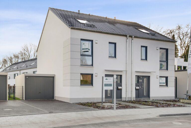 Außenansicht einer modernen Doppelhaushälfte in Duisburg Borkhofer Straße – helle Putzfassade, anthrazitfarbene Fenster und Haustür, Dachgaube mit Dachfenstern, angebaute Garage und gepflasterter Stellplatz, Neubau als Musterhaus