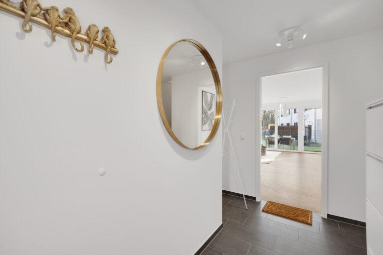 Heller Eingangsbereich mit dunkelgrauem Fliesenboden, rundem Wandspiegel mit goldenem Rahmen und dekorativer Garderobenhaken in Affenform, Blick in lichtdurchflutetes Wohnzimmer mit Gartenausblick – Immobilie in Duisburg